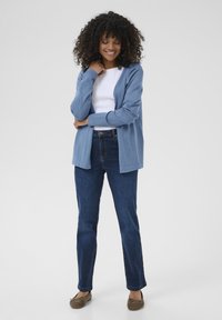 Cardigan azzurro chiaro realizzato in materiale morbido, indossato sopra una maglietta bianca, abbinato a jeans dritti blu scuro e scarpe slip-on marroni.