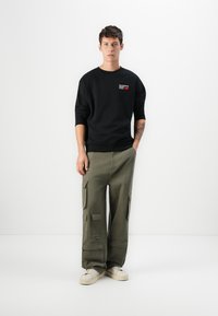 Black sweatshirt with logo, olive cargo pants with pockets, and white sneakers. Smooth texture, relaxed fit, and contrasting colors.