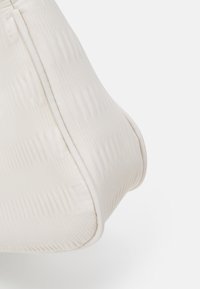 Beżowa poduszka o textured, falistej powierzchni, zaokrąglonych rogach i widocznym szwie wzdłuż krawędzi. Miękki materiał.