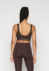 Sujetador deportivo marrón con tirantes anchos, espalda baja en forma de U y una banda gris contrastante. Combinado con leggings a juego de talle alto.
