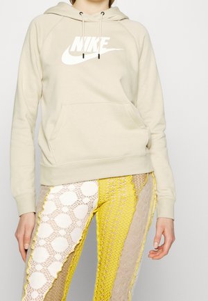 Beige Nike-Hoodie mit großem weißen Logo, ausgestattet mit einer Kängurutasche und einer Kapuze mit Zugband, kombiniert mit detailliert gemusterten gelben und beigen Hosen.