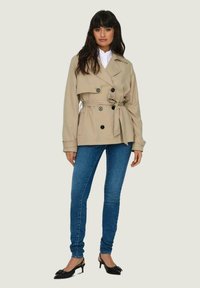 Cappotto trench beige doppiopetto con bottoni neri, cintura in vita e dettagli sulle spalle, abbinato a jeans skinny blu e tacchi neri.
