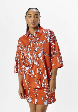 Camisa naranja de botones con mangas cortas, que presenta un patrón de hojas y flores en blanco. Tejido suave, ajuste holgado y detalle de bolsillo en la parte delantera.