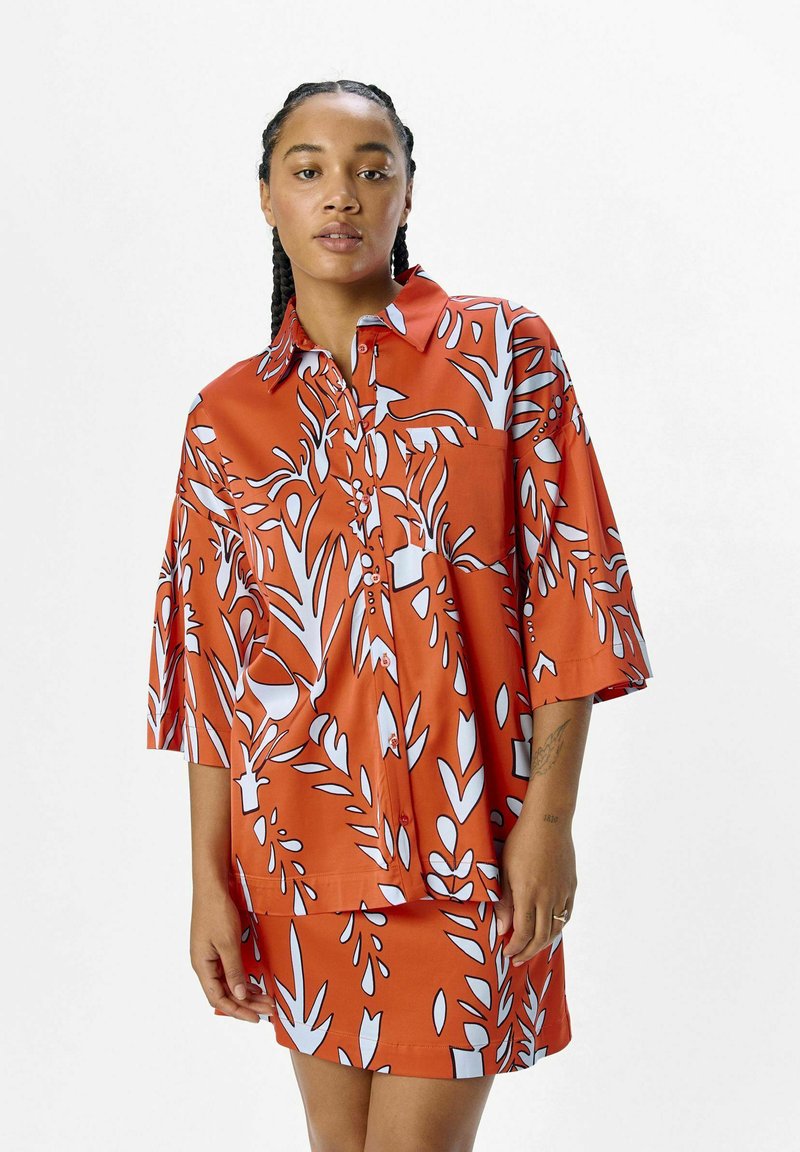 Camisa naranja de botones con mangas cortas, que presenta un patrón de hojas y flores en blanco. Tejido suave, ajuste holgado y detalle de bolsillo en la parte delantera.