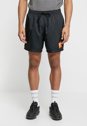 Mann trägt schwarze Shorts mit Kordelzug und einem orangefarbenen Logo-Patch, weiße Socken mit grauen Streifen, schwarze Sportschuhe, steht auf einem weißen Fußboden.
