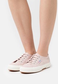 Pieds portant des baskets en toile à lacets rose clair avec des semelles épaisses blanches et une petite étiquette de logo sur le côté, sur fond blanc.