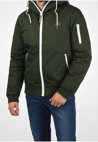 Giacca bomber verde scuro con zip bianca, tasche frontali e cappuccio con coulisse. Presenta un'etichetta con logo e polsini a righe.