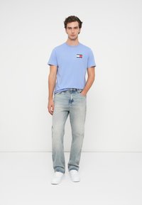 T-shirt de algodão azul claro com um pequeno emblema no peito, combinado com jeans azuis desbotados e tênis brancos, em pé sobre um fundo simples.