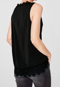 Černý top bez rukávů s texturovanou látkou, s krajkovým lemem na spodním okraji a kulatým výstřihem. Nesen s šedými džíny.
