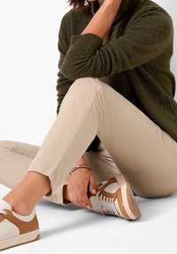 Olivgrön stickad cardigan över en mörk, tät skjorta, i kombination med beige slim-fit byxor och vita sneakers med bruna detaljer. Silverarmbandsdetalj.