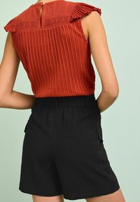 Top sin mangas de color óxido con detalle de volantes en el escote, combinado con pantalones cortos negros de cintura elástica que tienen bolsillos laterales.