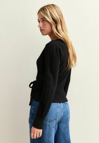 Svart stickad cardigan med knyt i midjan, långa ärmar och en figurnära design. Den mjuka texturen kompletterar de avslappnade blå denimjeansen.