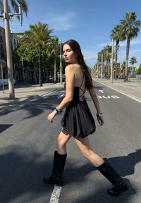 Schwarzes ärmelloses Kleid mit einem fließenden Rock, Rückendesign mit Überkreuzung; kombiniert mit hohen schwarzen Stiefeln mit dekorativen Beschlägen. Urbanes Straßenambiente.