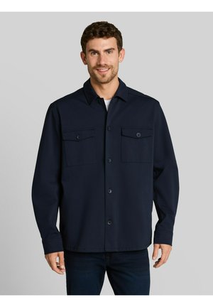 Uomo con capelli corti e ricci che sorride, indossa una giacca camicia blu scuro con due tasche sul petto e pantaloni scuri.