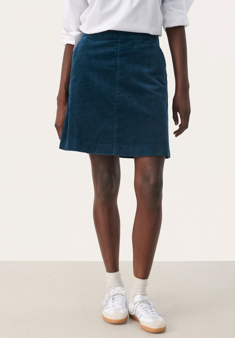 Jupe en velours côtelé d'un bleu canard profond avec une silhouette évasée, présentant des côtes verticales et une taille sans couture, associée à des baskets blanches et des chaussettes.