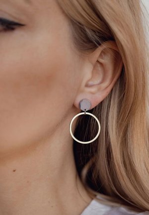 Orecchini a cerchio in oro con un perno circolare grigio opaco, caratterizzati da una superficie liscia e un design minimalista, appesi al lobo dell'orecchio.