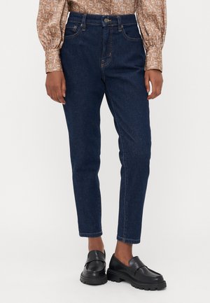 Mørkeblå denimjeans med smal passform, middels høyde på livet, frontlommer og subtile sømmer. Kombinert med svarte slip-on sko.