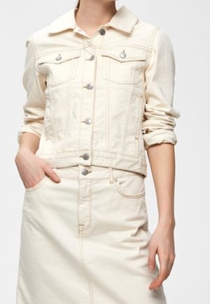 Femme portant une veste en jean crème aux manches retroussées et une jupe taille haute assortie, une main dans la poche, sur un fond uni.