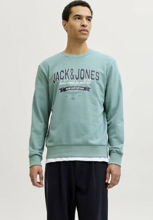 Hombre con sudadera de color verde claro de Jack & Jones y pantalones negros, de pie contra un fondo claro liso, mirando hacia la derecha.