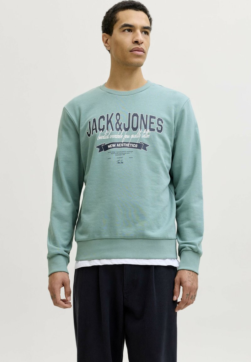 Hombre con sudadera de color verde claro de Jack & Jones y pantalones negros, de pie contra un fondo claro liso, mirando hacia la derecha.