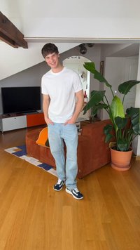 Jaunas vyras baltu marškinėliu ir šviesiomis džinsinėmis kelnėmis stovintis moderniame svetainės kambaryje su medine grindų danga, ruda sofa, dideliu žaliu augalu ir televizoriumi.