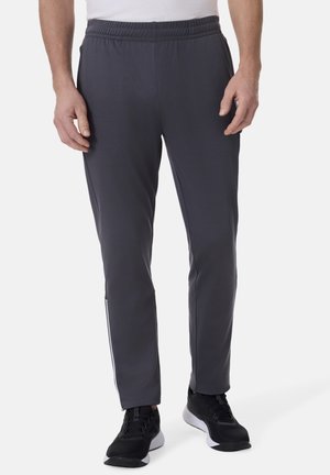 Mann trägt dunkelgraue Sporthose mit elastischem Bund und schwarze Turnschuhe, steht vor einem weißen Hintergrund.