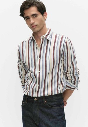 Ung mand iført en langærmet, knaplukket, stribet skjorte med op-rullede ærmer, stoppet ned i mørke jeans, stående mod en ensfarvet baggrund.