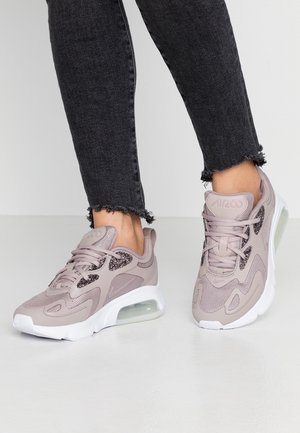 Osoba nosząca jasnoróżowe sportowe sneakersy z białymi podeszwami i detalem poduszki powietrznej, zestawione z czarnymi dżinsami z postrzępionym wykończeniem.