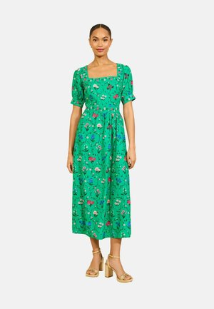Kobieta w zielonej sukience midi z kwadratowym dekoltem, bufkami na rękawach i wielobarwnym kwiatowym wzorem, zestawiona z beżowymi sandałami na klocku.