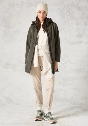 Chaqueta impermeable verde oliva, suéter crema anudado en la cintura, joggers beige y zapatillas verdes con acentos beige y gris.