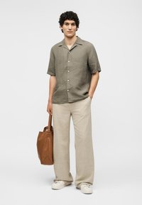 Άνδρας που φοράει ελιά κοντομάνικο πουκάμισο με κουμπιά, μπεζ φαρδιά παντελόνια, λευκά αθλητικά παπούτσια, κρατώντας καφέ δερμάτινη τσάντα tote.