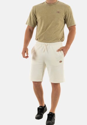 Camiseta de manga corta verde olivo con un pequeño logo; pantalones cortos color crema con cintura elástica y bolsillos laterales; zapatillas negras.