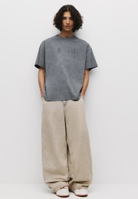 Grå oversized t-shirt med prægede bogstaver, beige brede bukser og hvide sneakers med brune detaljer. Minimalistisk design og blød tekstur.
