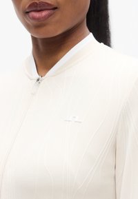 Μπεζ ζακέτα με φερμουάρ, με διακριτική κυματιστή υφή και λευκό λογότυπο ως θέμα. Διαθέτει ψηλό γιακά και ελαφρύ ύφασμα.