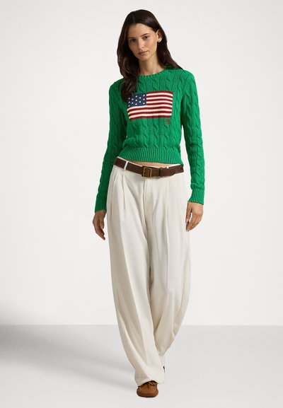 Camisola verde de malha com padrão de trança, com um gráfico da bandeira dos EUA no peito, combinada com calças brancas largas e sapatos castanhos.