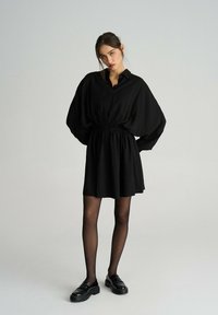 Czarna sukienka koszulowa z kołnierzykiem zapinanym na guziki, z wcięciem w talii, objętościowymi rękawami i kończąca się powyżej kolana. Połączona z czarnymi mokasynami.