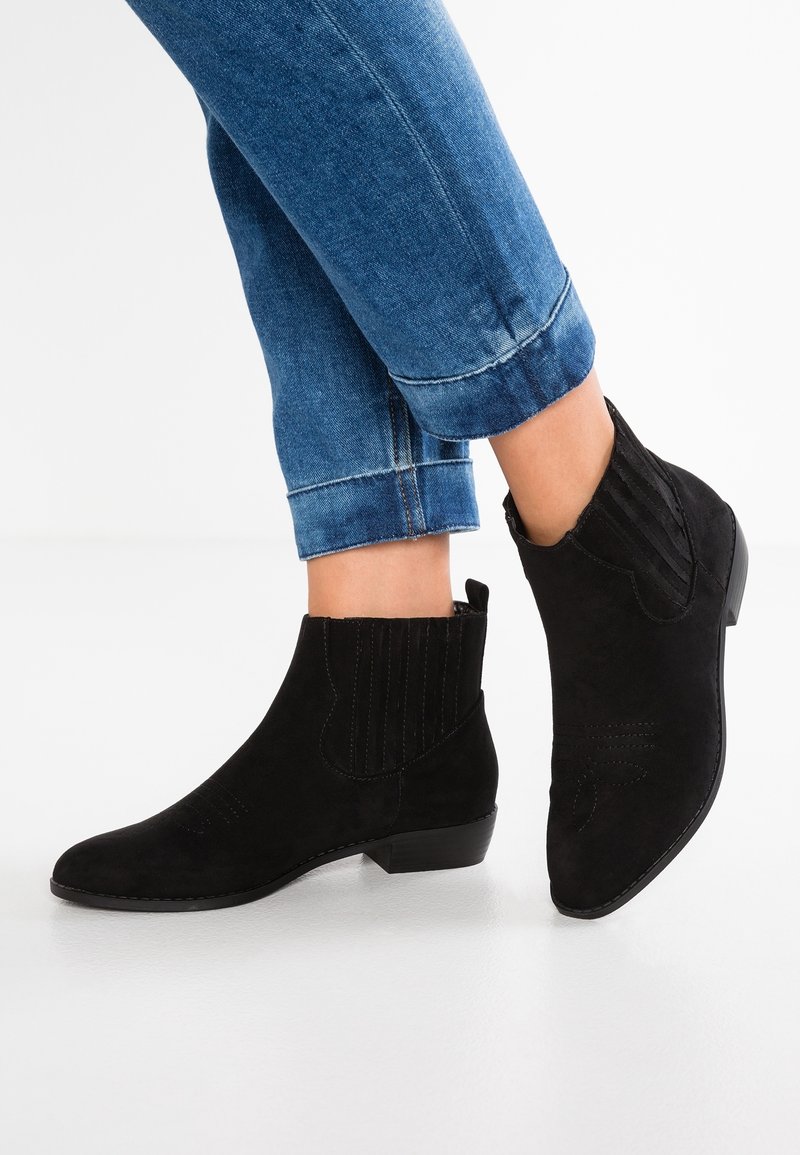 Svarta mocka ankelskor med låg klack, ribbad elastisk sida och sömnadsdetaljer. Bärs med uppvikta blå jeans.