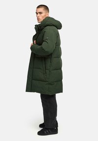 Piumino verde con cappuccio, caratterizzato da vita cinchata, motivo trapuntato e una toppa con logo sul braccio sinistro. Indossato sopra pantaloni e scarpe nere.