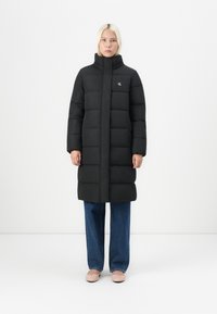 Svart polstret jakke med høy krage, frontlomme med glidelås, og CK-logo; kombinert med vide blå jeans og lyserosa ballerinasko.