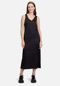 Schwarzes Satin-Midikleid mit V-Ausschnitt, ärmellosem Design und Seitenschlitzen. Kombiniert mit schwarzen Plateauschuhen für den Kontrast.
