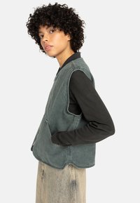 Groene denimvest met een ronde zoom en twee voorzakken, gecombineerd met een zwart shirt met lange mouwen en losse, lichtgekleurde spijkerbroeken.