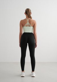 Top atletico verde chiaro con spalline incrociate, abbinato a leggings neri a coste e sneakers bianchi. Texture liscia e design aderente.