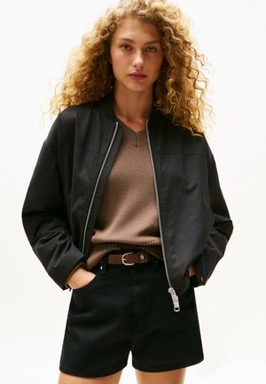 Chaqueta bomber negra con cremallera, suéter de punto marrón, pantalones cortos negros de cintura alta, cinturón marrón con hebilla dorada. Cabello rizado, fondo neutro.