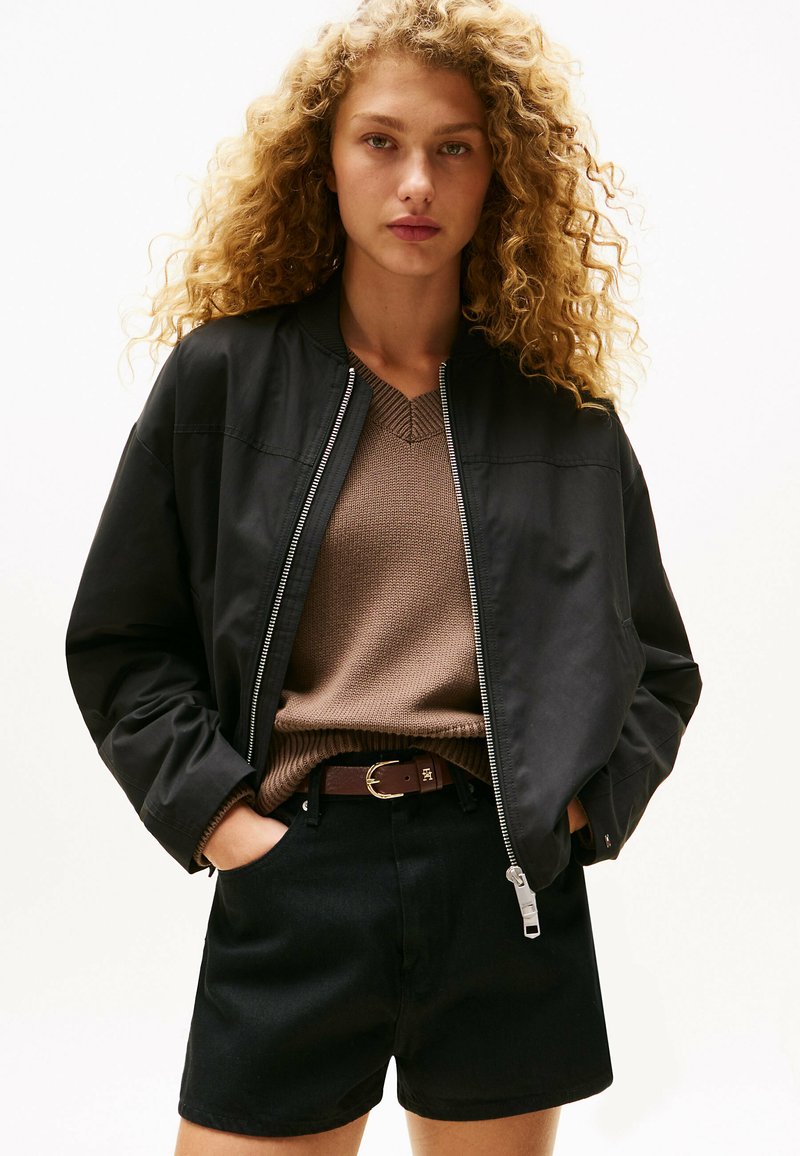 Czarna kurtka bomberka z zamkiem, brązowy sweter z dzianiny, czarne szorty z wysokim stanem, brązowy pasek z złotą klamrą. Kręcone włosy, neutralne tło.