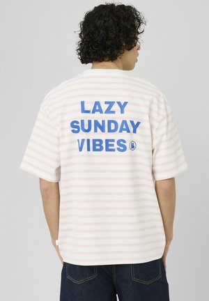 Personne aux cheveux bouclés portant un t-shirt rayé blanc et rose clair avec un texte bleu "LAZY SUNDAY VIBES" dans le dos, associée à un jean foncé.