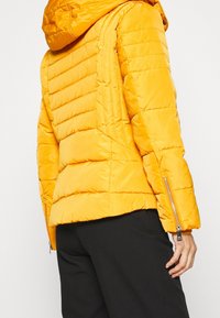 Giacca a vento in un vivace giallo, realizzata in materiale lucido con texture a trapezio, dotata di cappuccio e dettagli in zip argentata sulle maniche.