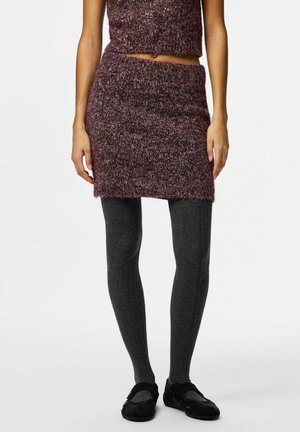 Frau trägt einen kastanienbraunen, gestrickten Rock und ein passendes bauchfreies Oberteil, dunkelgraue gemusterte Strumpfhosen und schwarze Slipper vor weißem Hintergrund.