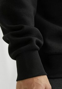 Tejido de sudadera negra con una superficie suave y texturizada. Las mangas con puños y el dobladillo tienen detalles acanalados para un ajuste ceñido.