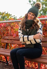 Bonnet tricoté vert foncé avec un pompon en fourrure, écharpe assortie et un pull crème à rayures noires. Assis sur un banc en bois.
