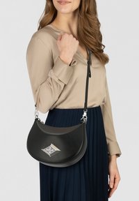 Sac bandoulière en cuir noir avec un design courbé, une quincaillerie argentée et un logo en forme de losange. Sangle réglable et texture lisse.